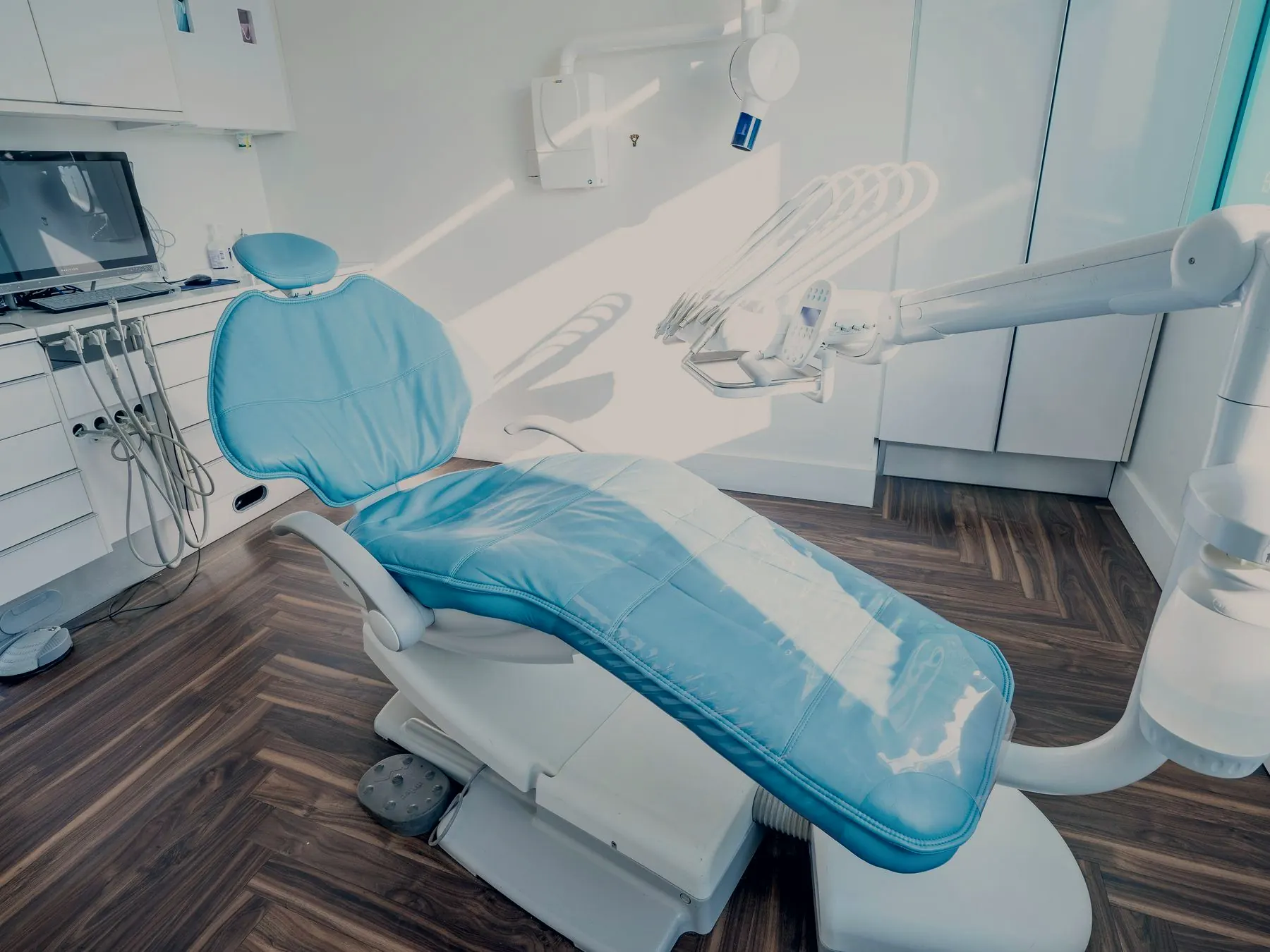 Dental treatment room with a reclined chair, overhead lamp and a wall-mounted imaging display at the adjacent workstation