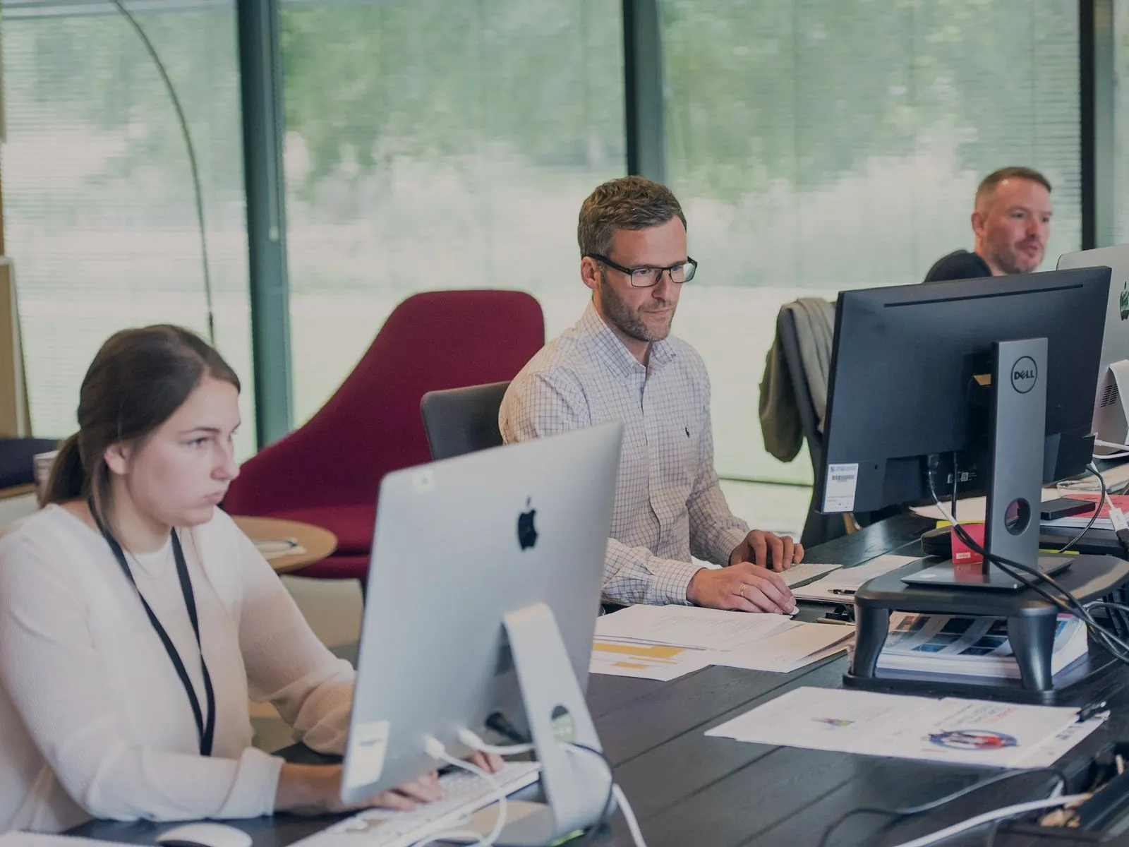 Three people working at desktop computers along a shared desk in a daylit open-plan office