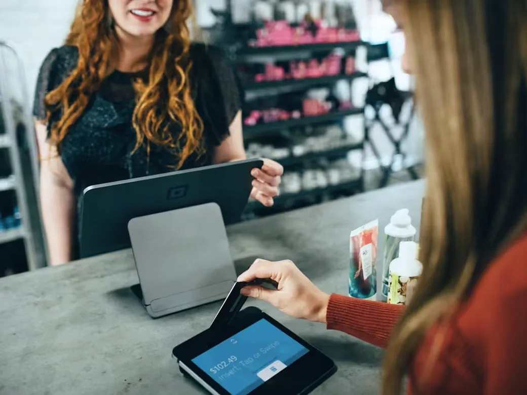 A customer paying by card at a small retail counter while the shop assistant looks on beside a tablet point-of-sale stand