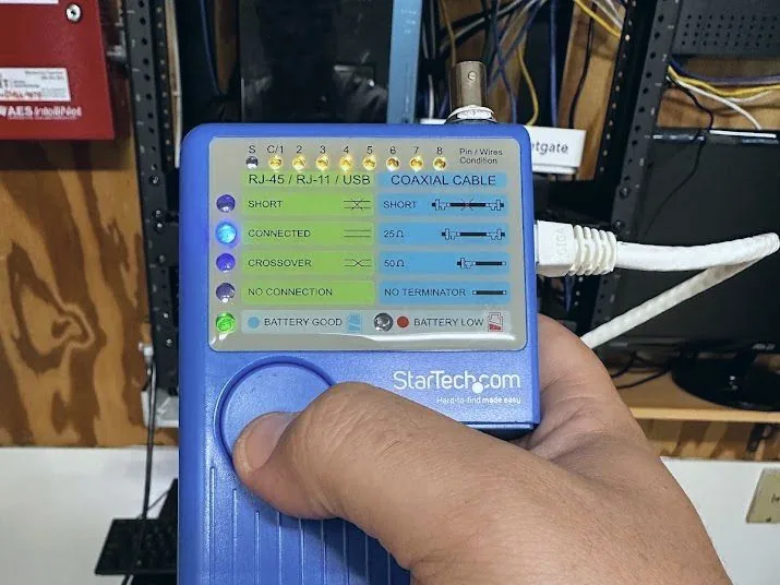 Hand holding a cable tester displaying a lit CONNECTED indicator, with a patch cable attached and an equipment rack behind