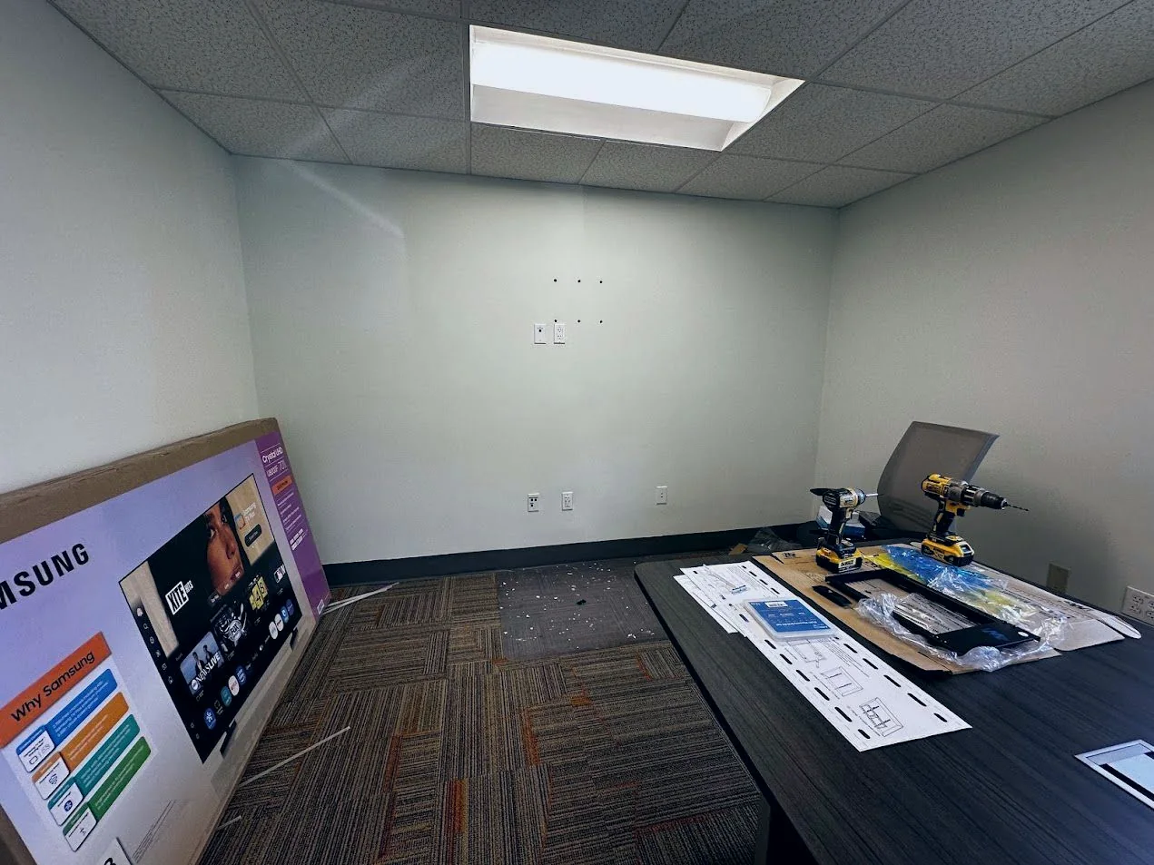 A conference room part way through an audio-visual fit-out, with cable outlets in the wall, mounting hardware and cordless drills on the table and a boxed display leaning against the wall
