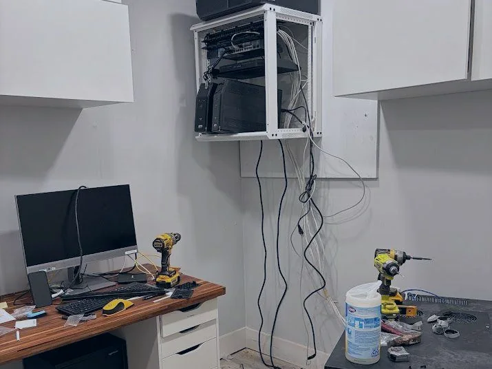 A wall-mounted rack part-built with its door open, network cabling run down the wall, and cordless drills on the bench below