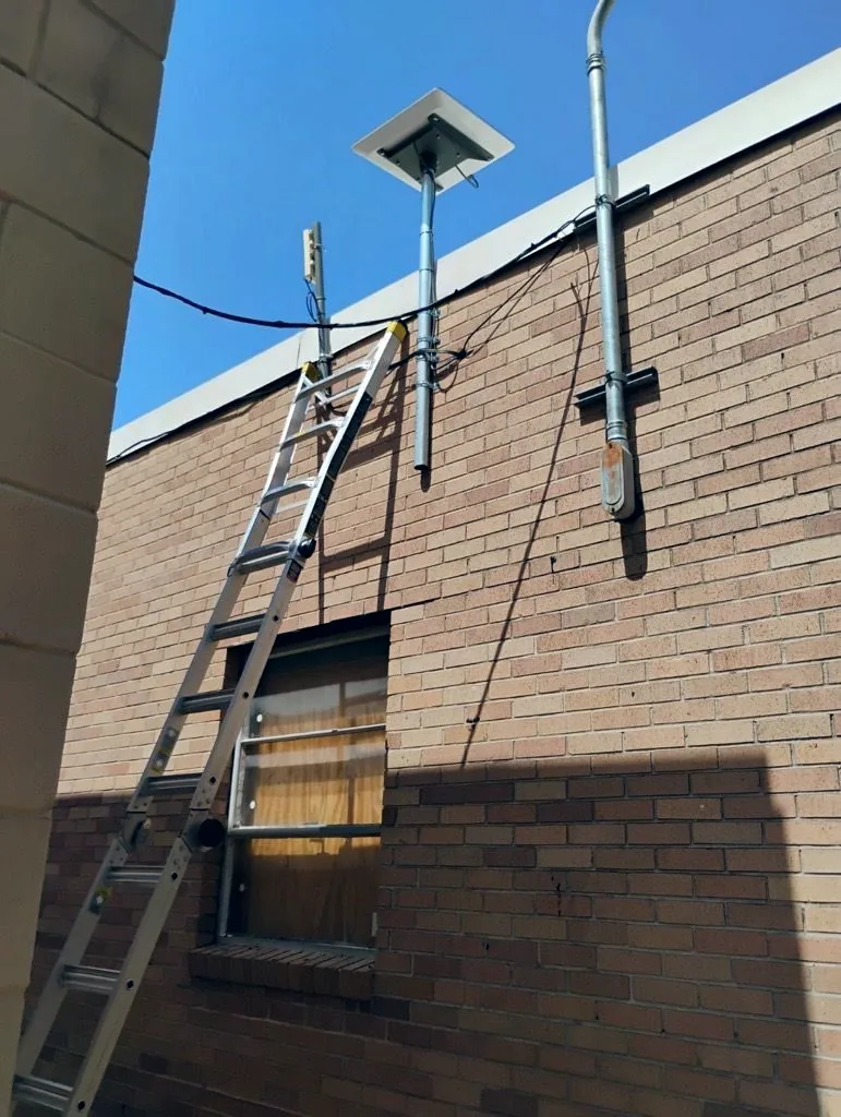 Extension ladder against a brick exterior wall below a satellite dish on a mast, with cabling routed down to a conduit