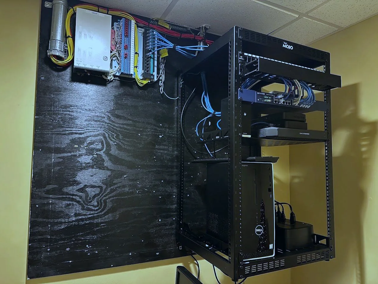 Finished IT room with an open-frame rack of network equipment beside a plywood backboard carrying punchdown blocks and dressed telephone cabling