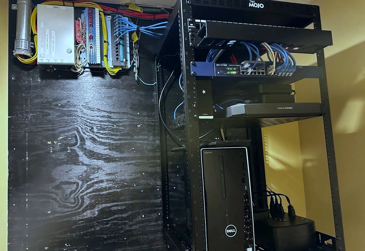 Open-frame equipment rack holding a network switch, business modem and desktop computer, beside a plywood backboard with punchdown blocks and telephone cabling