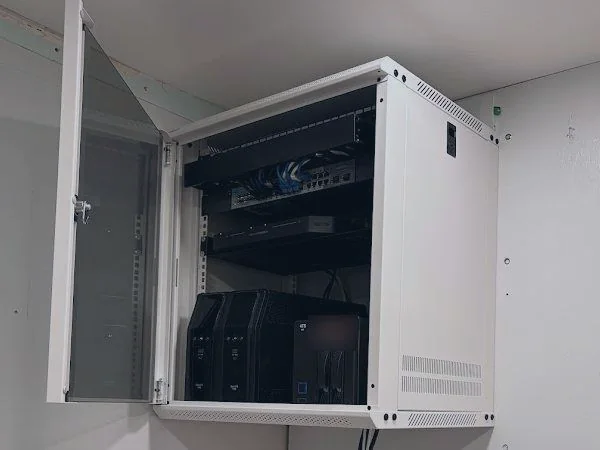 A wall-mounted rack with its door open showing a patch panel, network switch and storage units, with cabling dressed down the wall