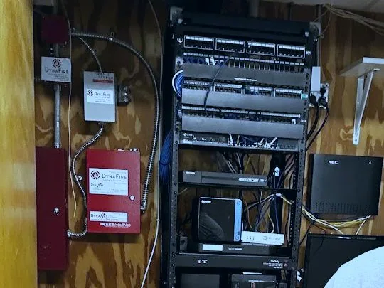 Wall-mounted equipment rack with patch panels, a network switch, backup power and a phone system, beside a fire alarm panel