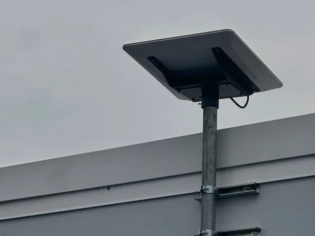 Satellite dish mounted on a galvanised mast above a building parapet, braced to the wall with standoff brackets and a weatherproof junction box