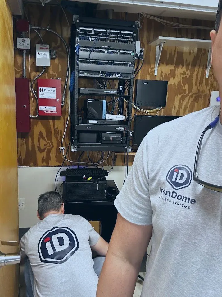 Two technicians in IronDome shirts working at a wall-mounted equipment rack with patch panels, a switch and a phone system