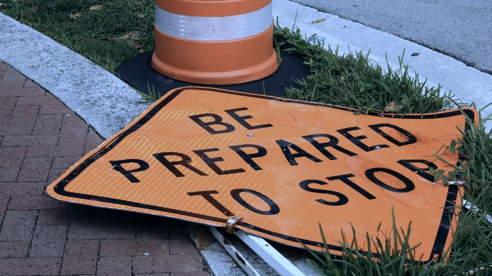A traffic drum and a folded orange BE PREPARED TO STOP sign lying on a grass verge beside a brick footpath